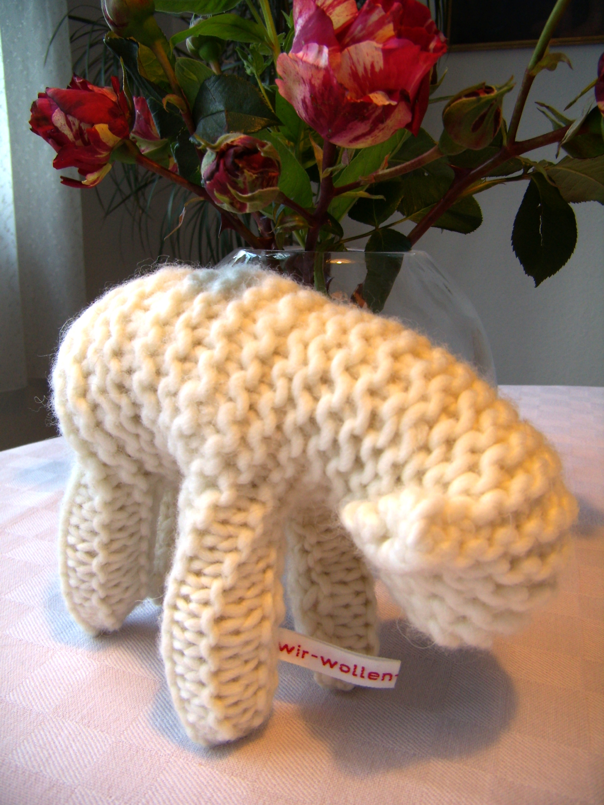 Gestricktes Lamm aus weißer Wolle mit Wäschebändchen "Wir wollen Vielfalt" vor einer Vase mit roten Rosen.