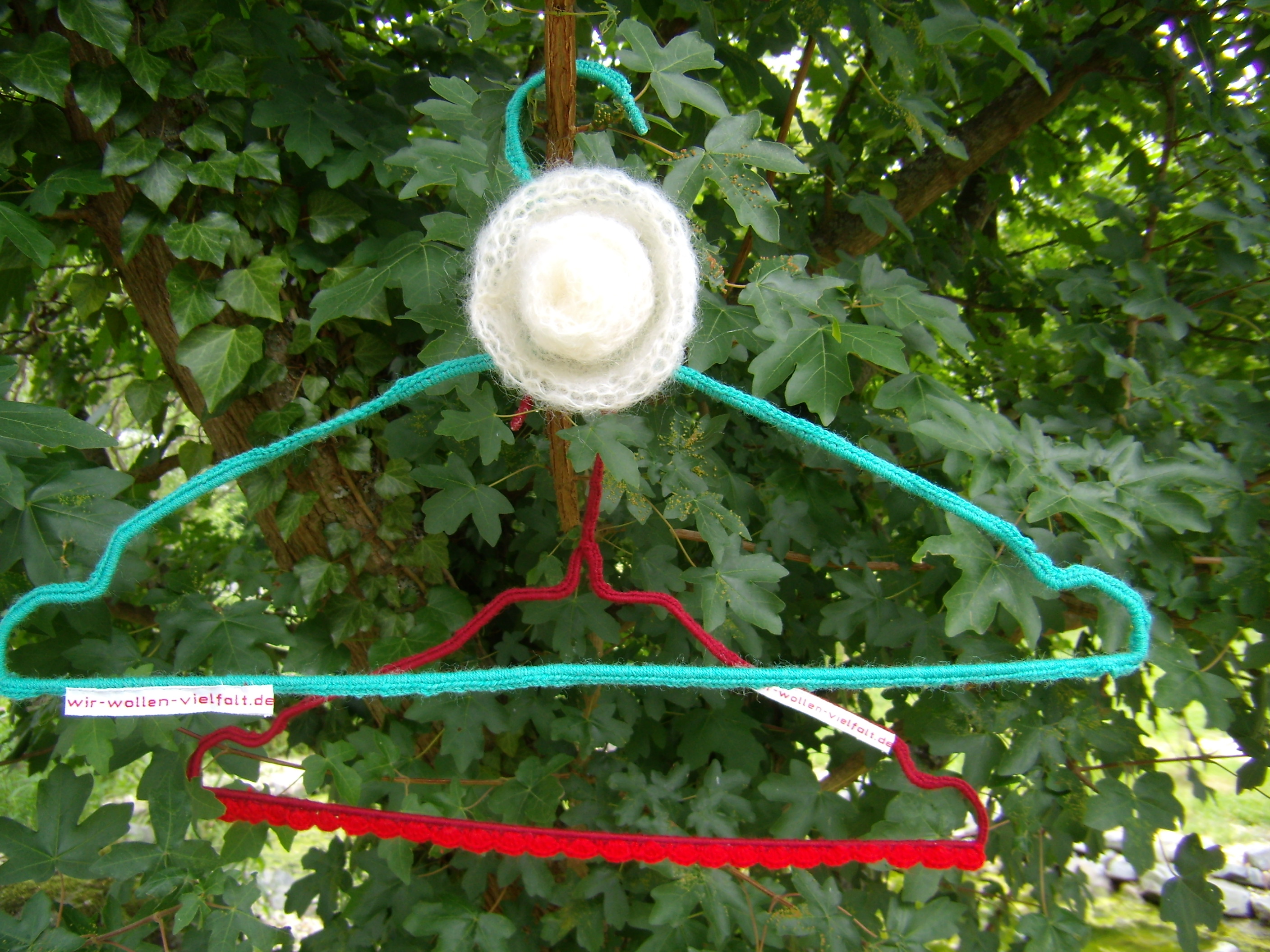Zwei Draht-Kleiderbügel mit der Aufschift "wir-wollen-vielfalt.de", der eine rot, der andere grün umhäkelt, hängen in einem Busch voller Laub.