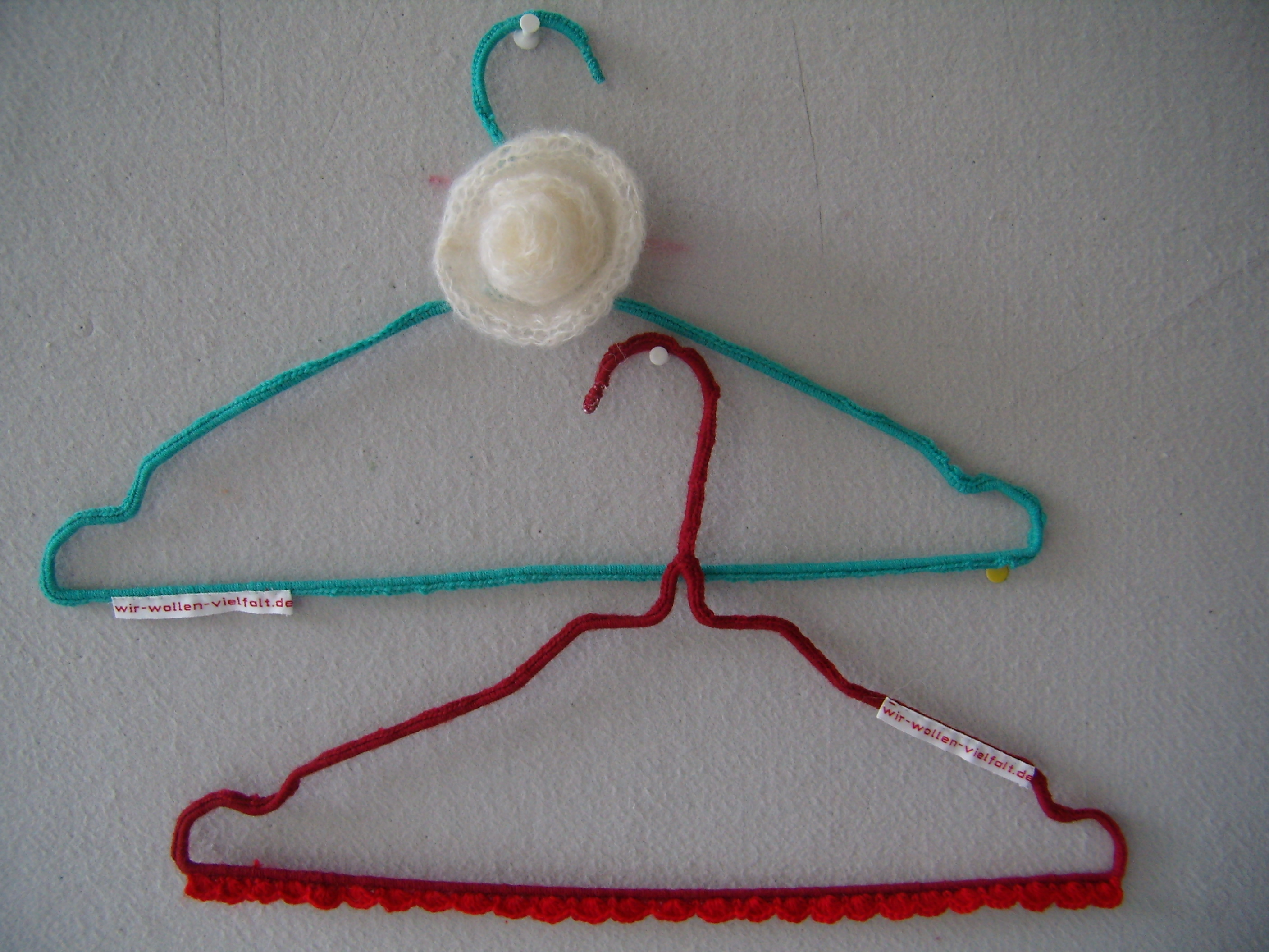 Zwei Draht-Kleiderbügel mit der Aufschift "wir-wollen-vielfalt.de", der eine rot, der andere grün umhäkelt, hängen an einer hellgrauen Pinwand.