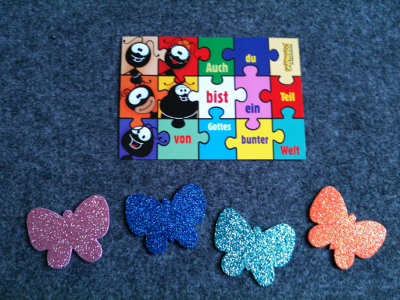 Vier Moosgummi-Schmetterlinge und eine Postkarte mit der Aufschrift: "Auch du bist ein Teil von Gottes bunter Welt."