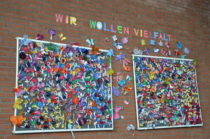 Ziegelwand mit zwei Pinwänden voller Vielfalter, darüber in Papierbuchstaben die Worte: "Wir wollen Vielfalt".