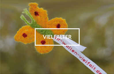 Gelber Häkel-Schmetterling mit grünem Körper und Wäscheband mit Aufschrift "wir-wollen-vielfalt.de". In der Mitte des Bildes steht das Wort "Vielfalter".